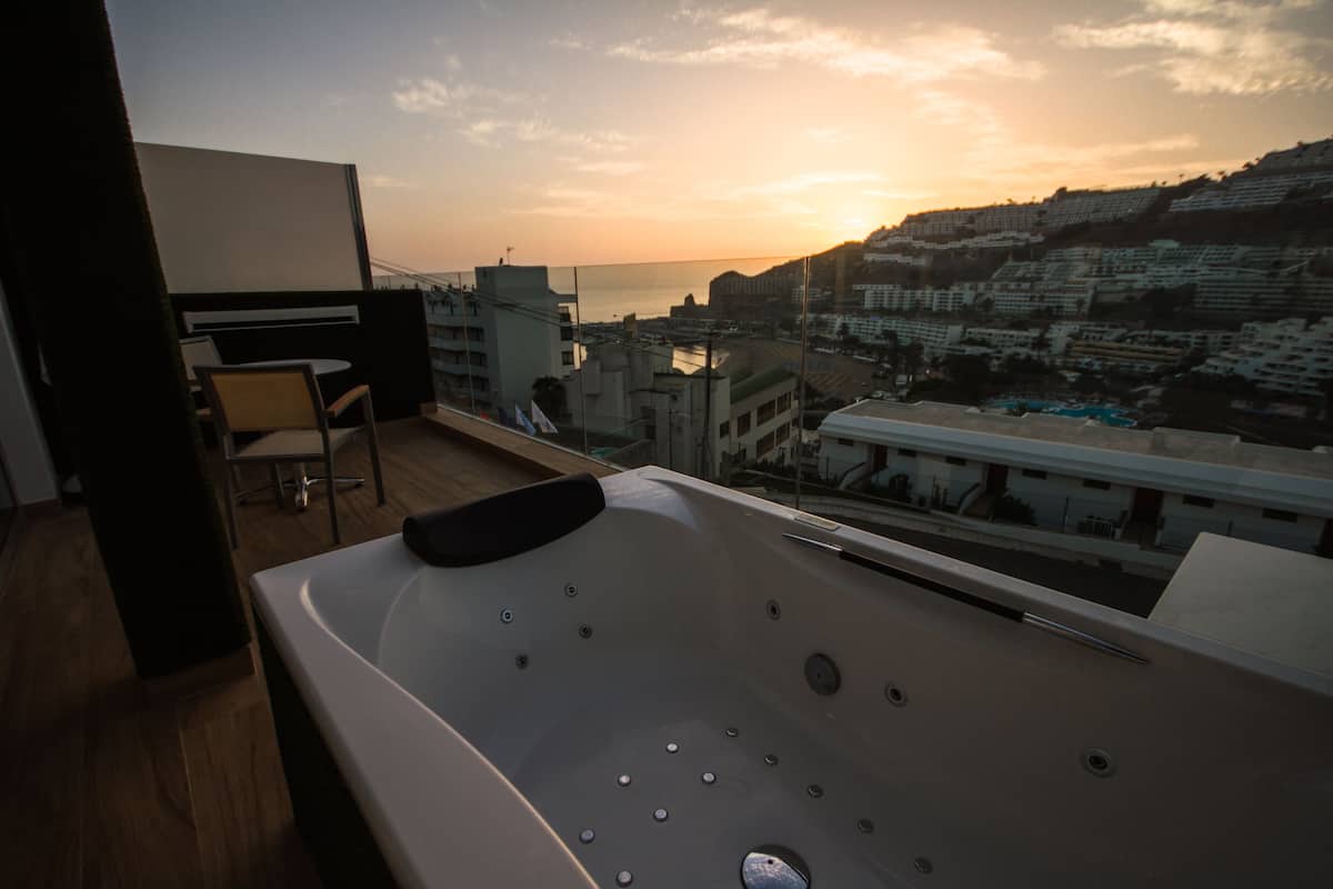 Un baño moderno con una bañera grande, una silla y vistas a un paisaje urbano al atardecer.