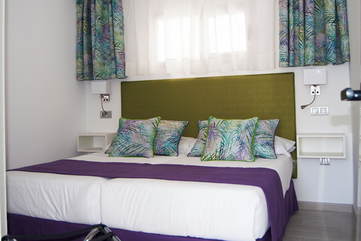 Una habitación de hotel con una cama, cabecera, mesita de noche y una ventana con cortinas.