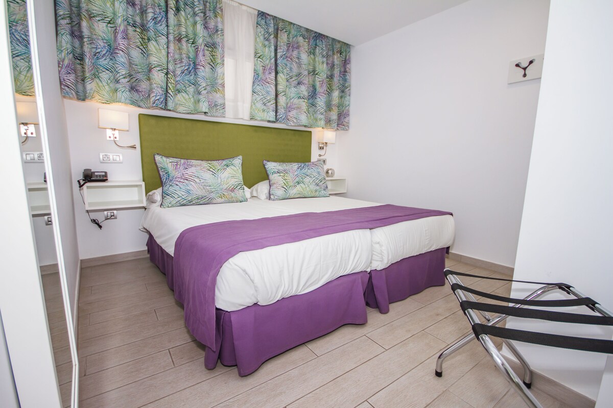 Un dormitorio con cama, cabecera, mesita de noche, lámpara y perchero de pared.