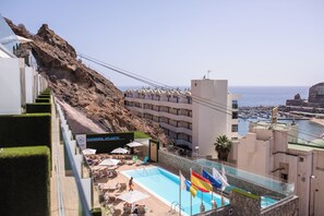 Un hotel con zona de piscina, mobiliario exterior y vistas al mar.