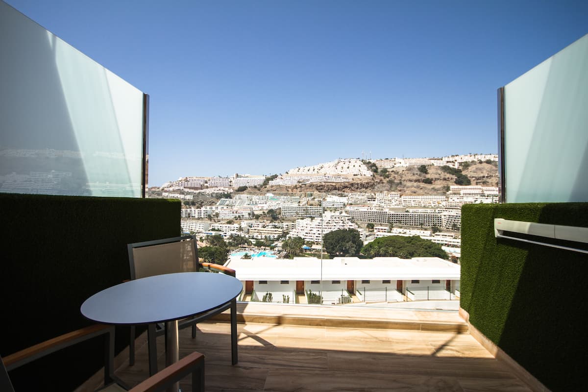 Un balcón con mesa y sillas, con vista a un paisaje urbano de edificios y zonas verdes.