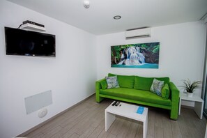 Una habitación con un sofá verde, un televisor de pantalla plana montado en la pared, un cuadro de una cascada y una mesita con controles remotos.