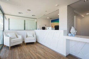 Un área de recepción con un mostrador blanco, dos sillones tapizados, una computadora portátil y un cuadro de una playa.