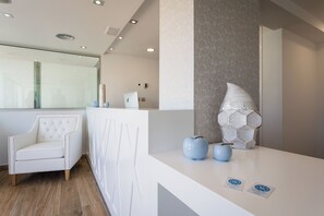 Un área de recepción con un mostrador blanco, un sillón blanco con capitoné y un elemento decorativo hexagonal en la pared.