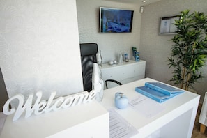 Una recepción con un cartel de 'Welcome', un televisor montado en la pared, un armario con varios objetos y una planta en maceta.
