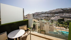 Un balcón con vistas a un pueblo costero, una piscina y un área de comedor con sillas y una mesa.
