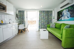 A modern living room with a green sofa, a white coffee table, and a white chair. There is a kitchen area with white cabinets and a microwave. A painting of a waterfall is on the wall. - IG Nachosol Atlantic & Yaizasol by Servatur (Mogan)