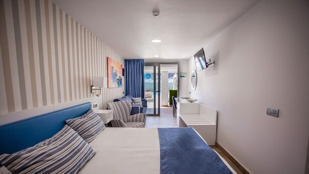 Habitación de hotel con cama, zona de estar con sofá y sillones, una mesita y baño con espejo y lavamanos.