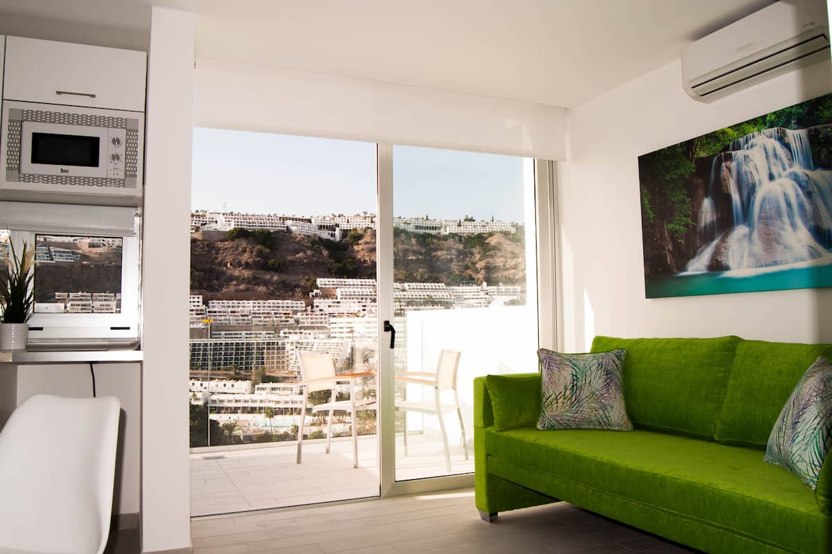 Una sala de estar moderna con un sofá verde, una zona de comedor con sillas, una cocina con microondas y un ventanal con vistas a la ciudad.