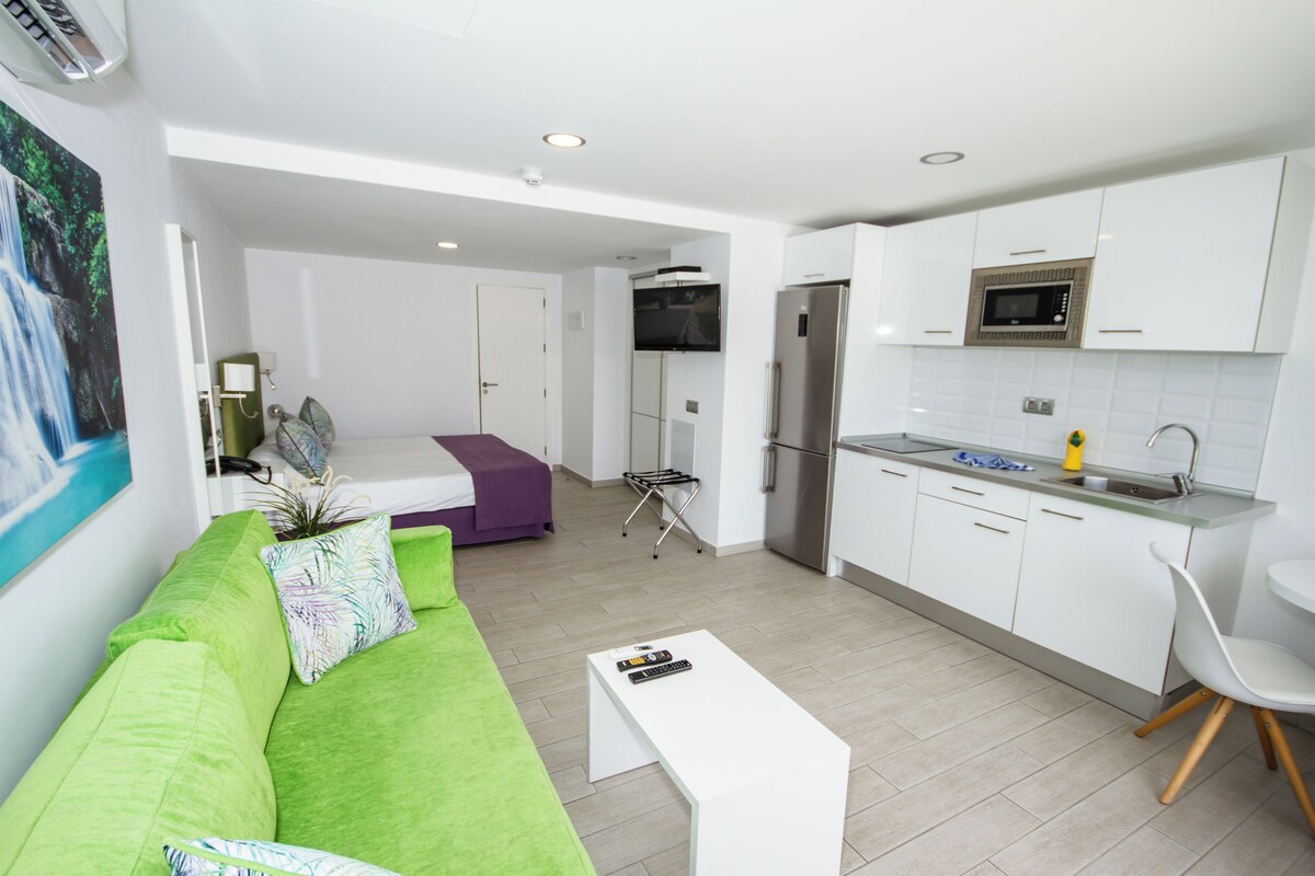 Un apartamento moderno con sala de estar que incluye un sofá verde, una mesa de centro blanca y un televisor. La cocina tiene gabinetes blancos, un refrigerador de acero inoxidable y un microondas.