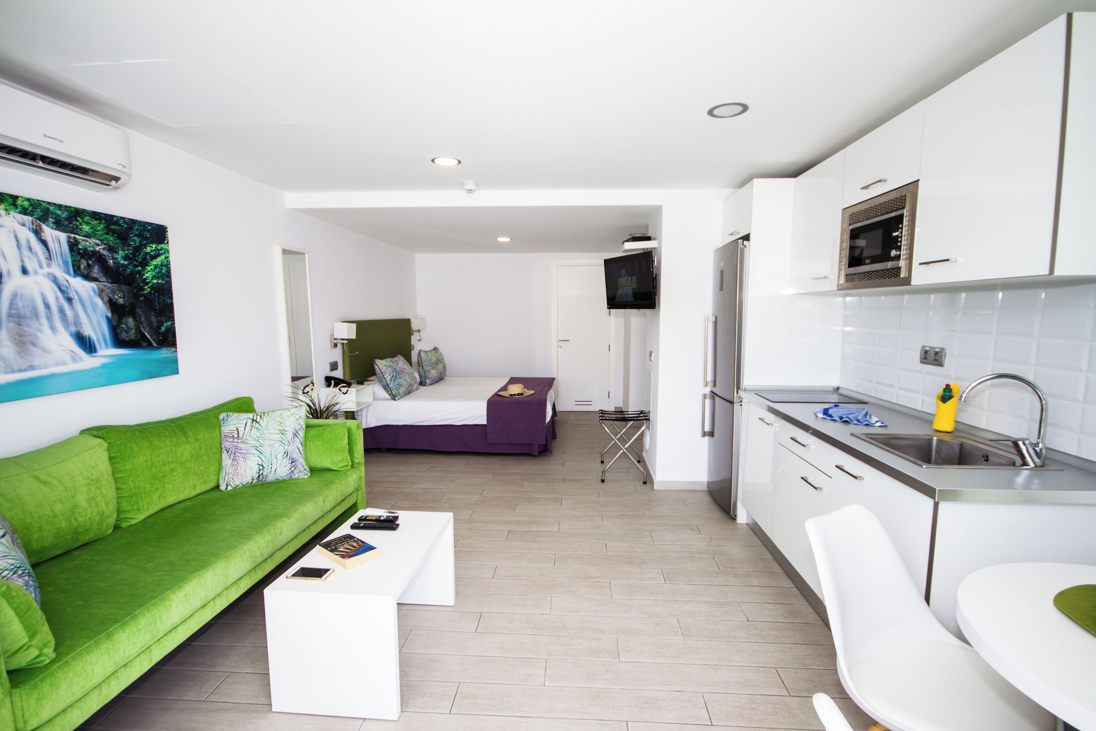 A modern hotel room with a green sofa, a white coffee table, a bed with a purple bedspread, a small TV, a kitchen area with a sink and a microwave, and a bathroom with a toilet.