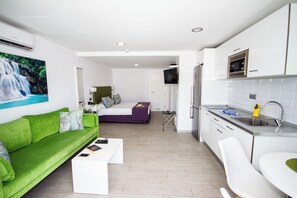 A modern hotel room with a green sofa, a white coffee table, a bed with a purple bedspread, a small TV, a kitchen area with a sink and a microwave, and a bathroom with a toilet. - IG Nachosol Atlantic & Yaizasol by Servatur (Mogan)