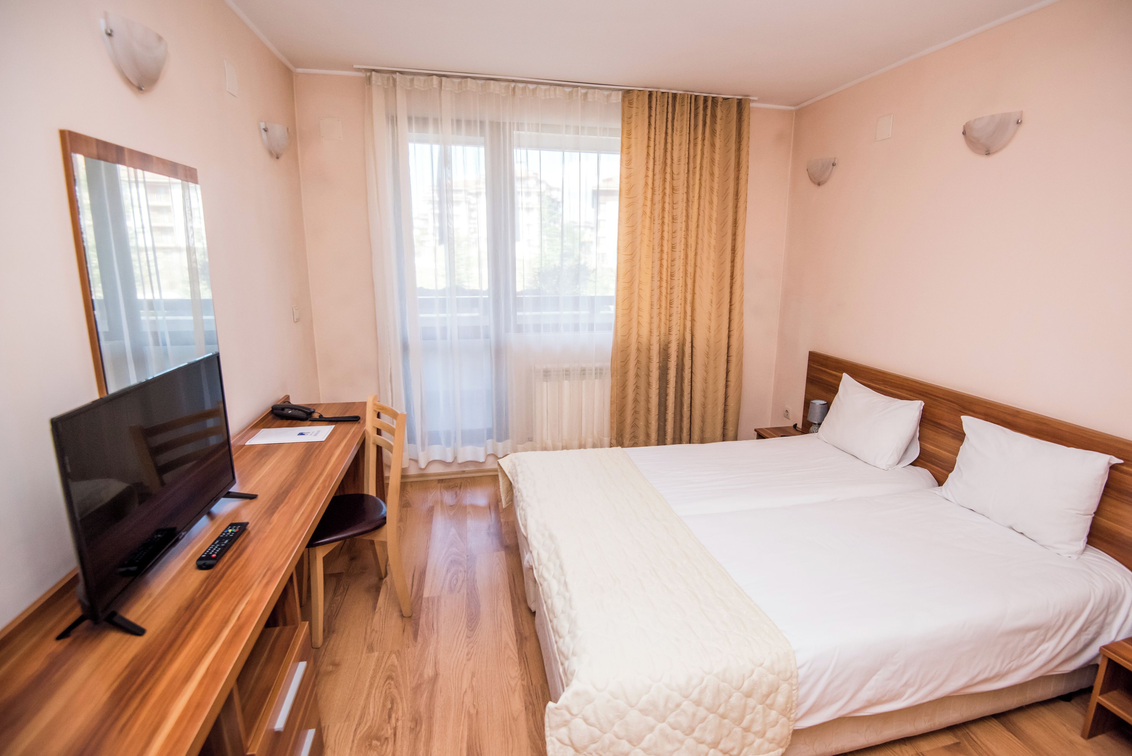 A hotel room with a wooden desk, a TV, a bed with white bedding, a chair, and a window with curtains.
