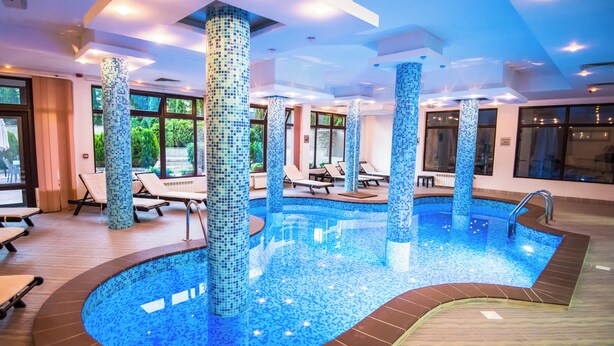 An indoor swimming pool with tiled walls and columns, surrounded by lounge chairs and a sunbed.