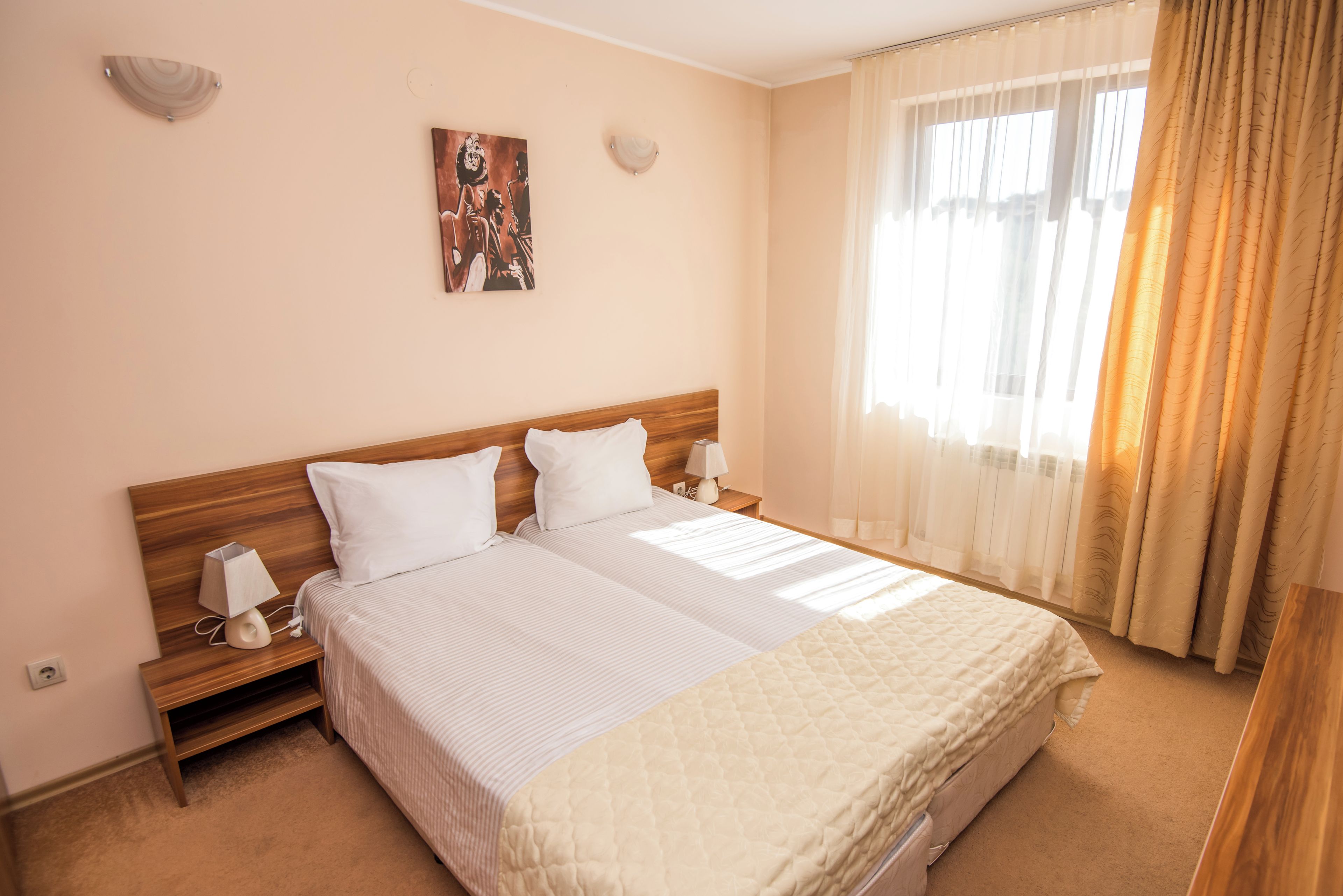 A hotel room with a large bed, two bedside tables, a nightstand, a wall-mounted lamp, a painting, and a window with curtains.