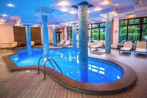 An indoor swimming pool with tiled columns, lounge chairs, and a view of greenery outside.
