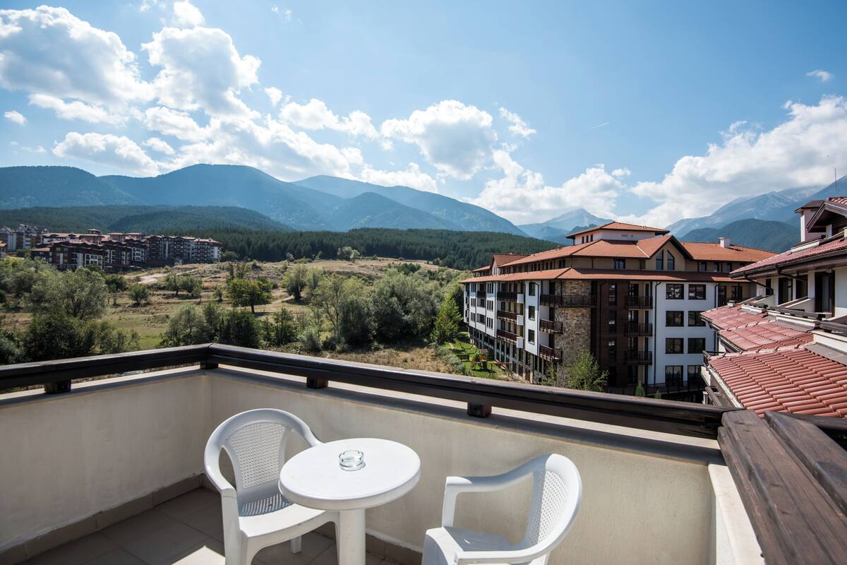 A balcony with two white chairs and a small round table, overlooking a mountainous landscape with buildings in the distance.
