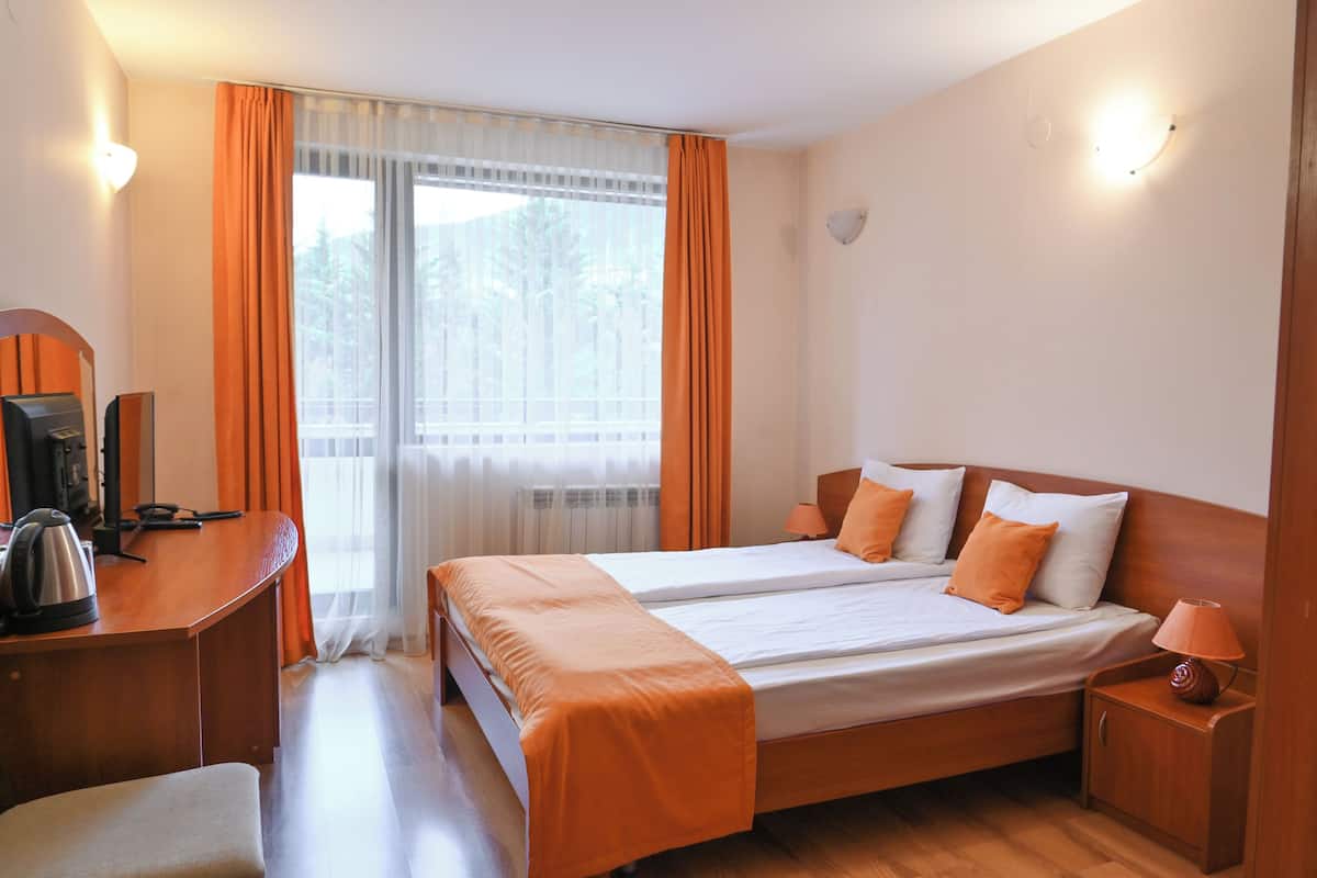 A hotel room with a double bed, a desk with a kettle, a chair, and a nightstand with a lamp.