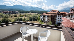 A balcony with two white plastic chairs and a small round table, overlooking a mountainous landscape with buildings in the background. - Orbilux (Bansko)