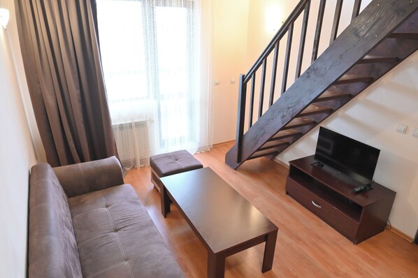 A living room with a sofa, a coffee table, a TV on a wooden stand, and a staircase. - Orbilux (Bansko)
