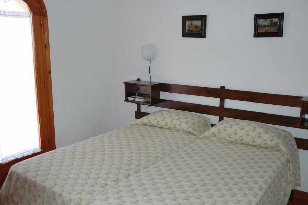 A bedroom with a wooden bed frame, two pillows, a bedside table with a lamp, and two framed pictures on the wall.