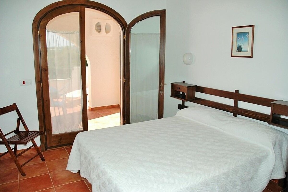 Une chambre à coucher comprenant un lit, une tête de lit en bois, une chaise, une armoire et une porte donnant sur un balcon.