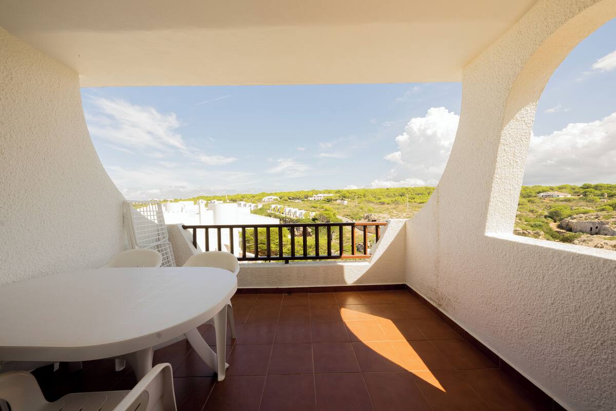A balcony with a white table and chairs, a view of greenery and buildings in the distance, and a clear sky.