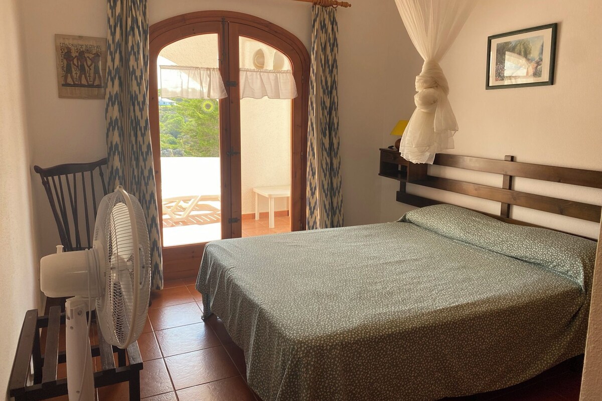 A bedroom with a bed, a chair, a fan, and a view of the outside.