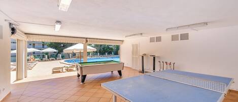 A room with a ping pong table, a pool table, and a view of an outdoor pool area.