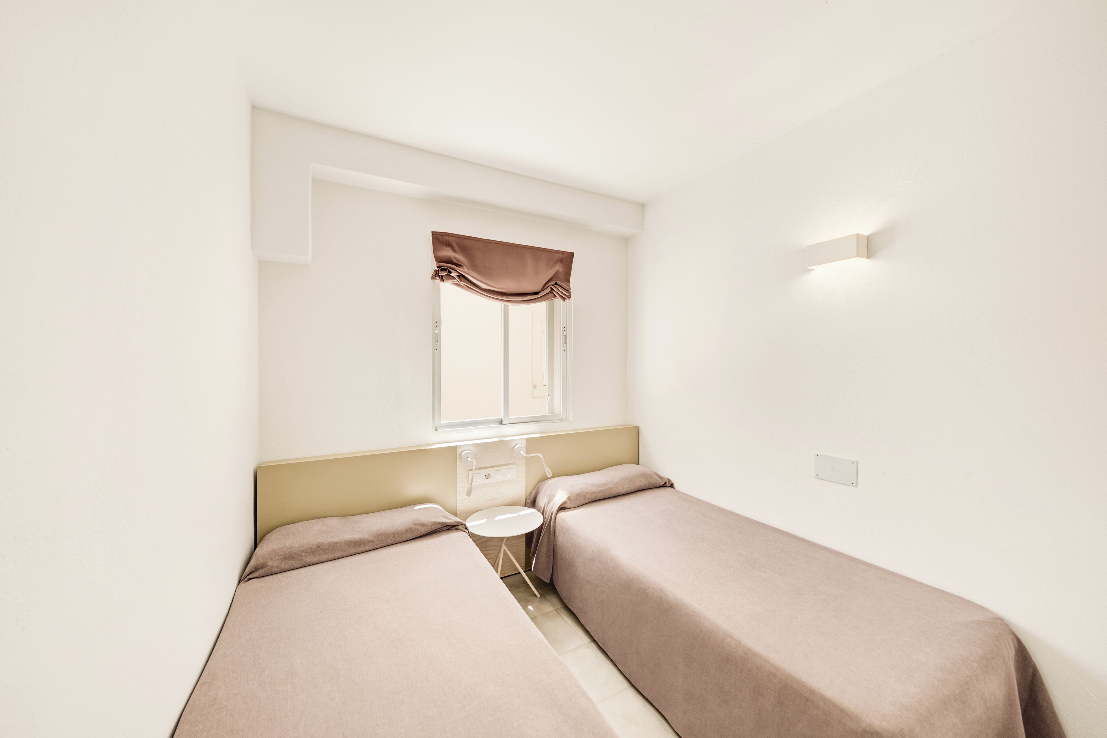 A room with two single beds, a small table, a window with curtains, and a wall-mounted light fixture.