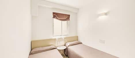 A room with two single beds, a small table, a window with curtains, and a wall-mounted light fixture.