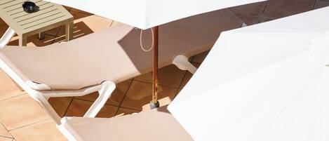 A sunlit outdoor seating area with white lounge chairs and a large white umbrella providing shade.