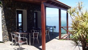 Un edificio de piedra con un patio que tiene mesas y sillas, una pérgola de madera y vista al océano.