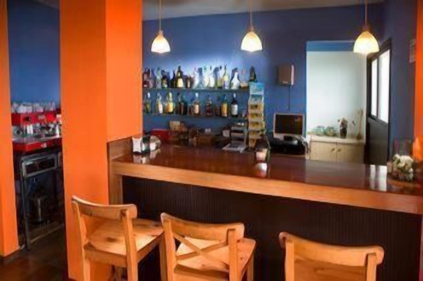 Zona de bar amb un taulell de fusta, tres seients de fusta i una estant amb ampolles.