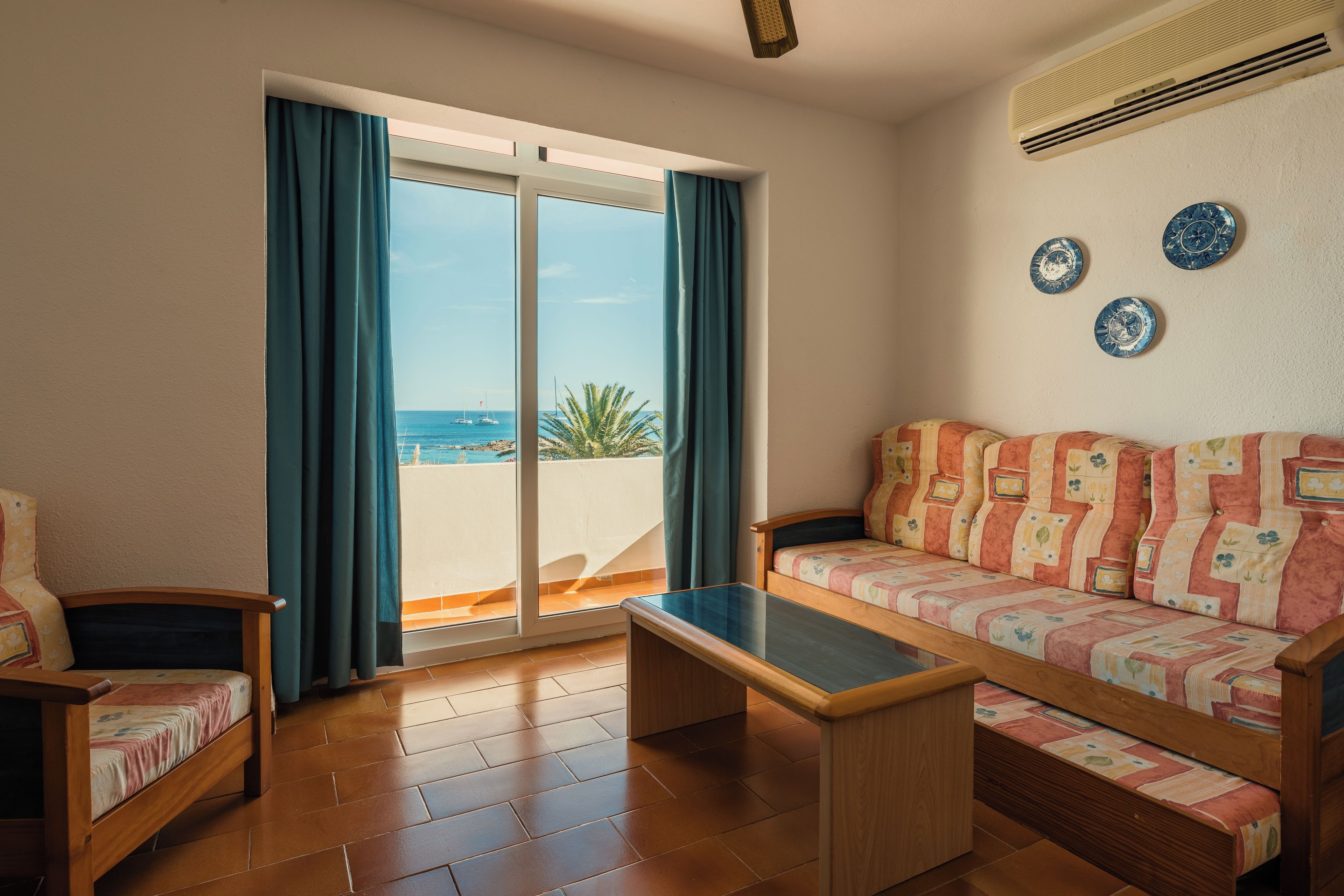 A room with a sofa, a small table, and a chair. There is a sliding glass door leading to a balcony with a view of the sea.