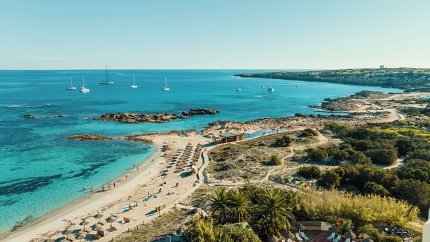 A coastal beach with clear turquoise waters, boats, and a rocky shoreline.