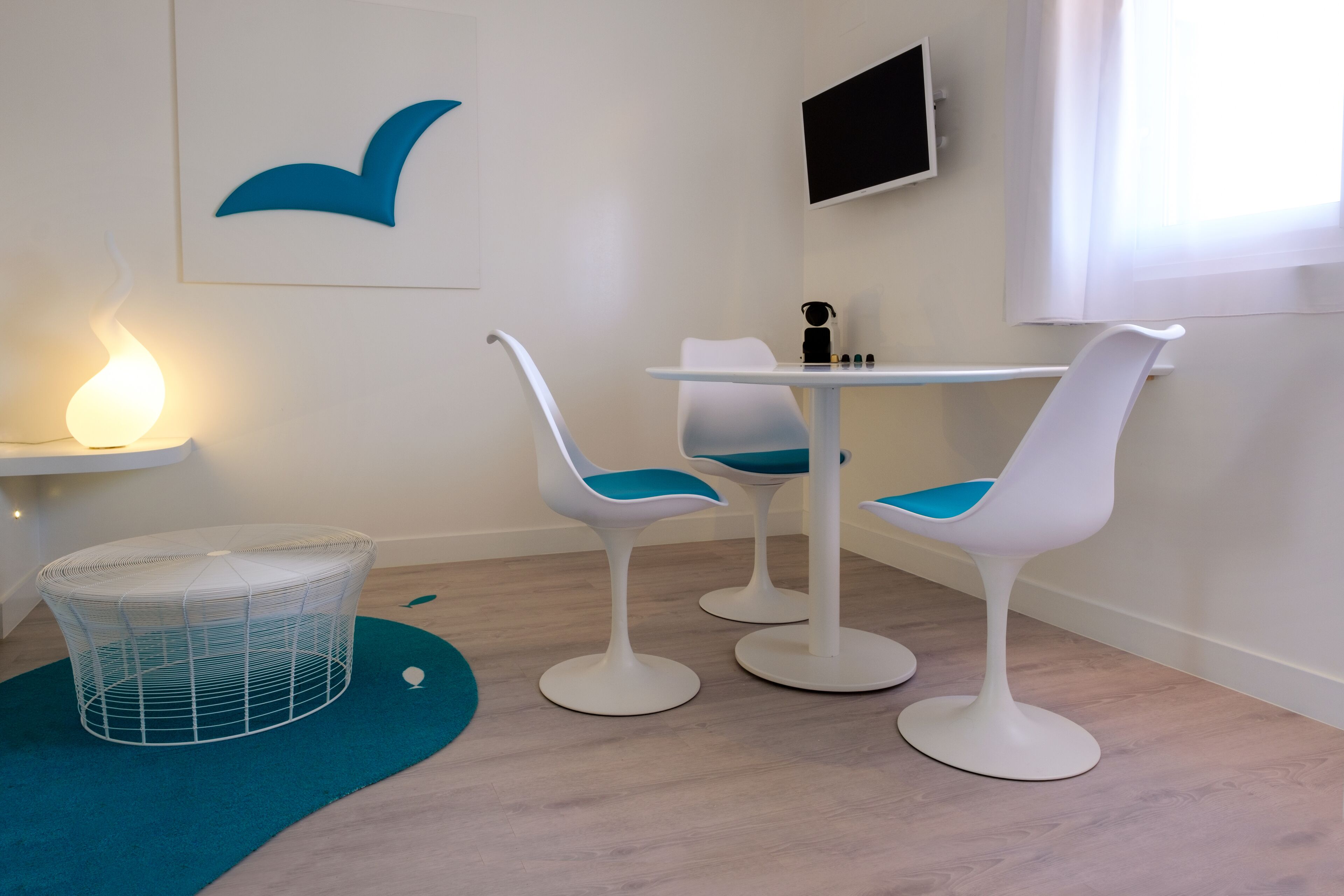 A room with a white wall, a blue whale mural, a white table with two chairs, a small white shelf with a lamp, and a white chair with a blue cushion.