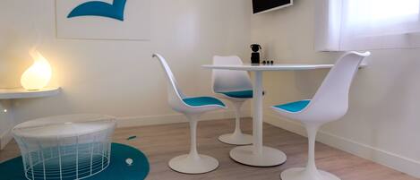 A room with a white wall, a blue whale mural, a white table with two chairs, a small white shelf with a lamp, and a white chair with a blue cushion.