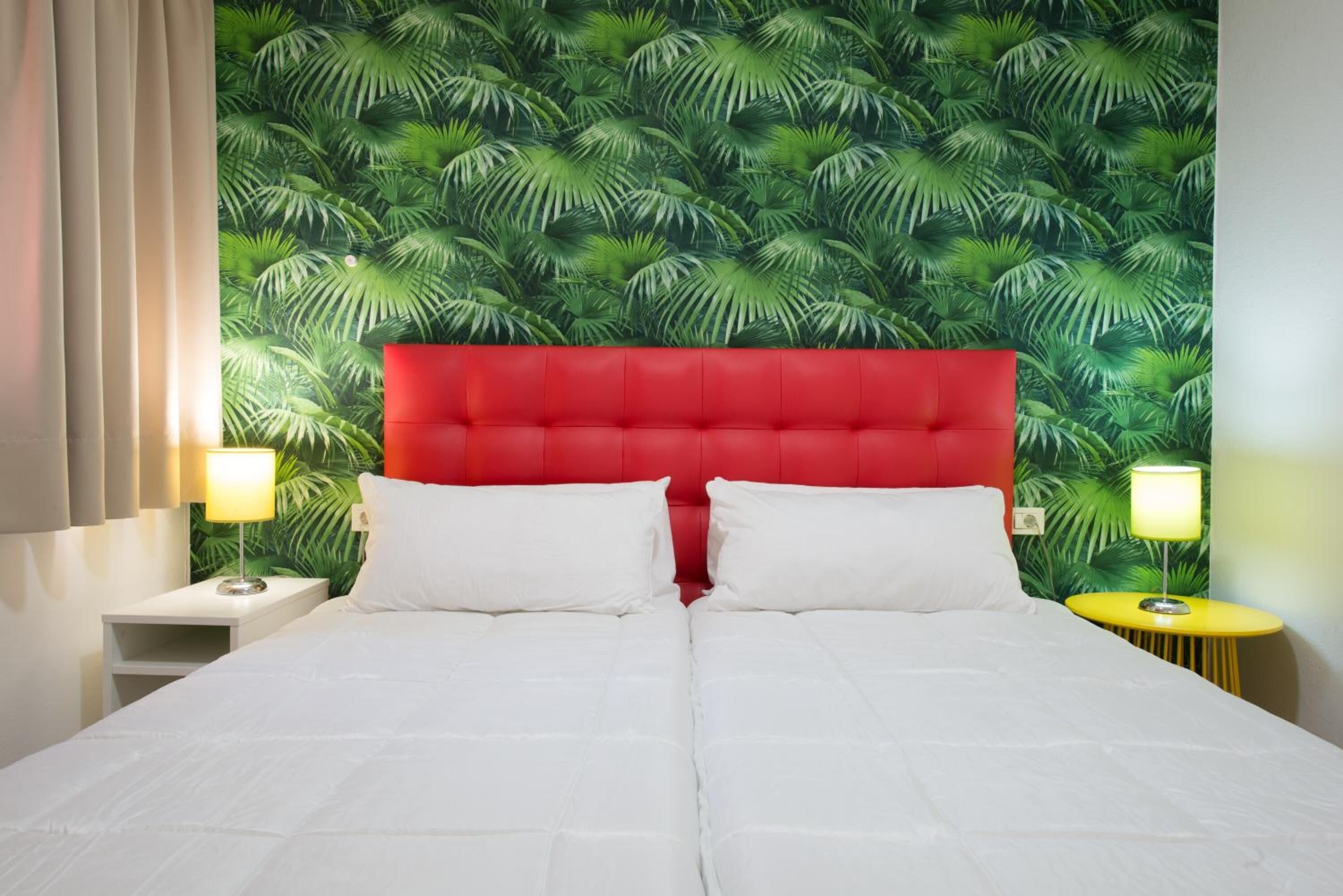 A hotel room with a red headboard, white bedding, and a tropical leaf wallpaper. There are two bedside tables with lamps.