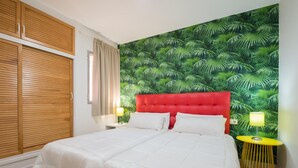 Une chambre à coucher avec une tête de lit rouge, du linge de lit blanc, un papier peint à motifs tropicaux et une table de chevet jaune.