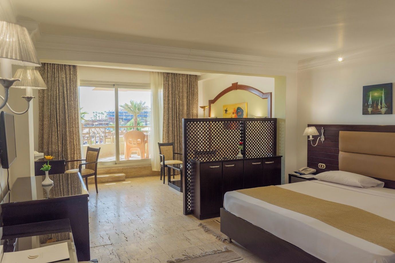 A hotel room with a bed, a desk with a lamp, a chair, a view of the outside, and a decorative partition.