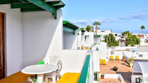 A balcony with a white table and chairs, a green railing, and a view of a white building with yellow chairs and potted plants. - Apartamentos Isabel (Tias)