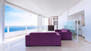 Een moderne woonkamer met een groot raam met uitzicht op zee, een paarse bank en een flatscreentelevisie.