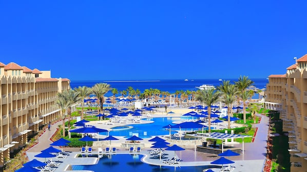 A resort with a central pool surrounded by lounge chairs and umbrellas, a clear view of the ocean, and a multi-story building with balconies.