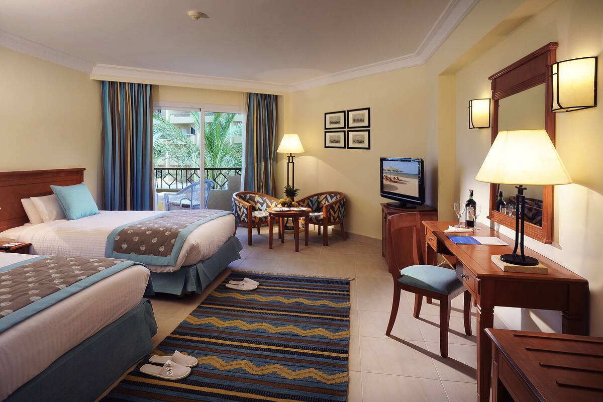 A hotel room with two beds, a desk, a chair, a TV, and a balcony with curtains.