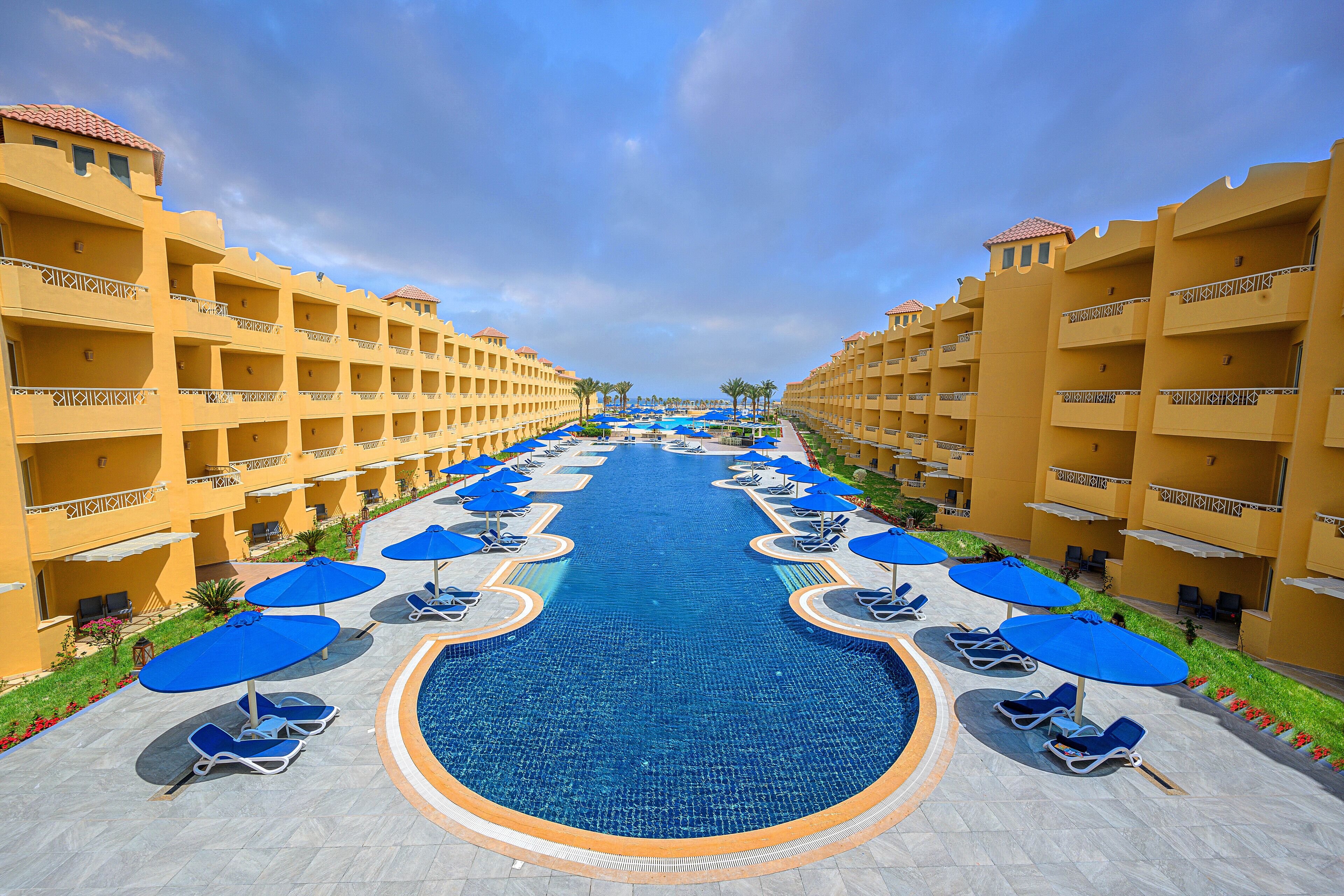 A hotel with a central swimming pool, multiple balconies, and sun loungers with umbrellas.