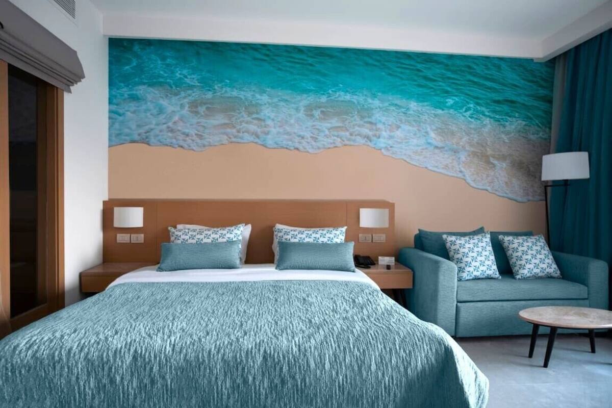 A hotel room with a large ocean-themed mural, a bed with a patterned bedspread, a sofa with matching cushions, and a small round table.