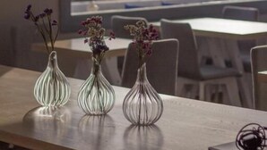 A dining area with a large window overlooking a body of water, a wooden table, and three glass vases with flowers. - Grotta Hotel (Naxos)