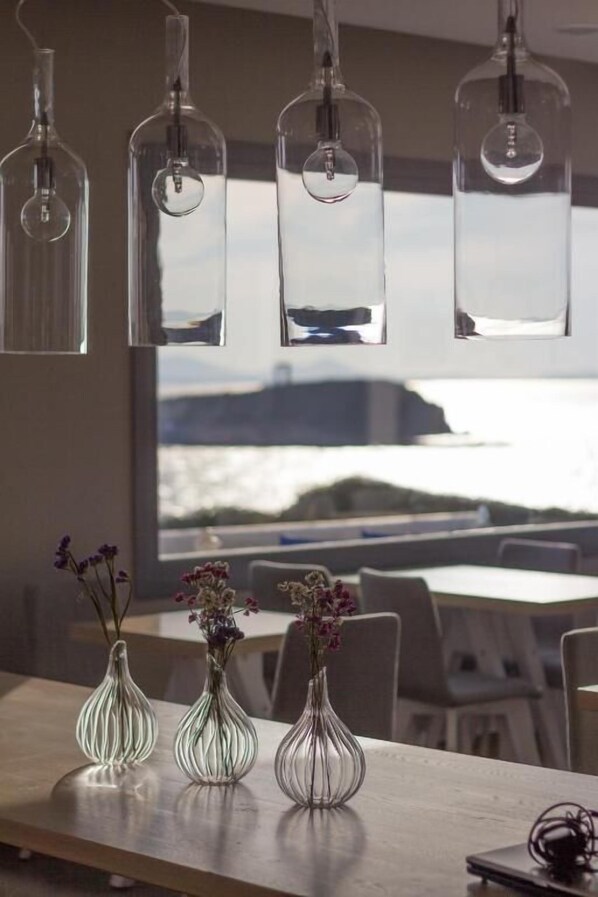 Un coin repas avec une grande fenêtre donnant sur un plan d’eau, une table en bois et trois vases en verre ornés de fleurs.