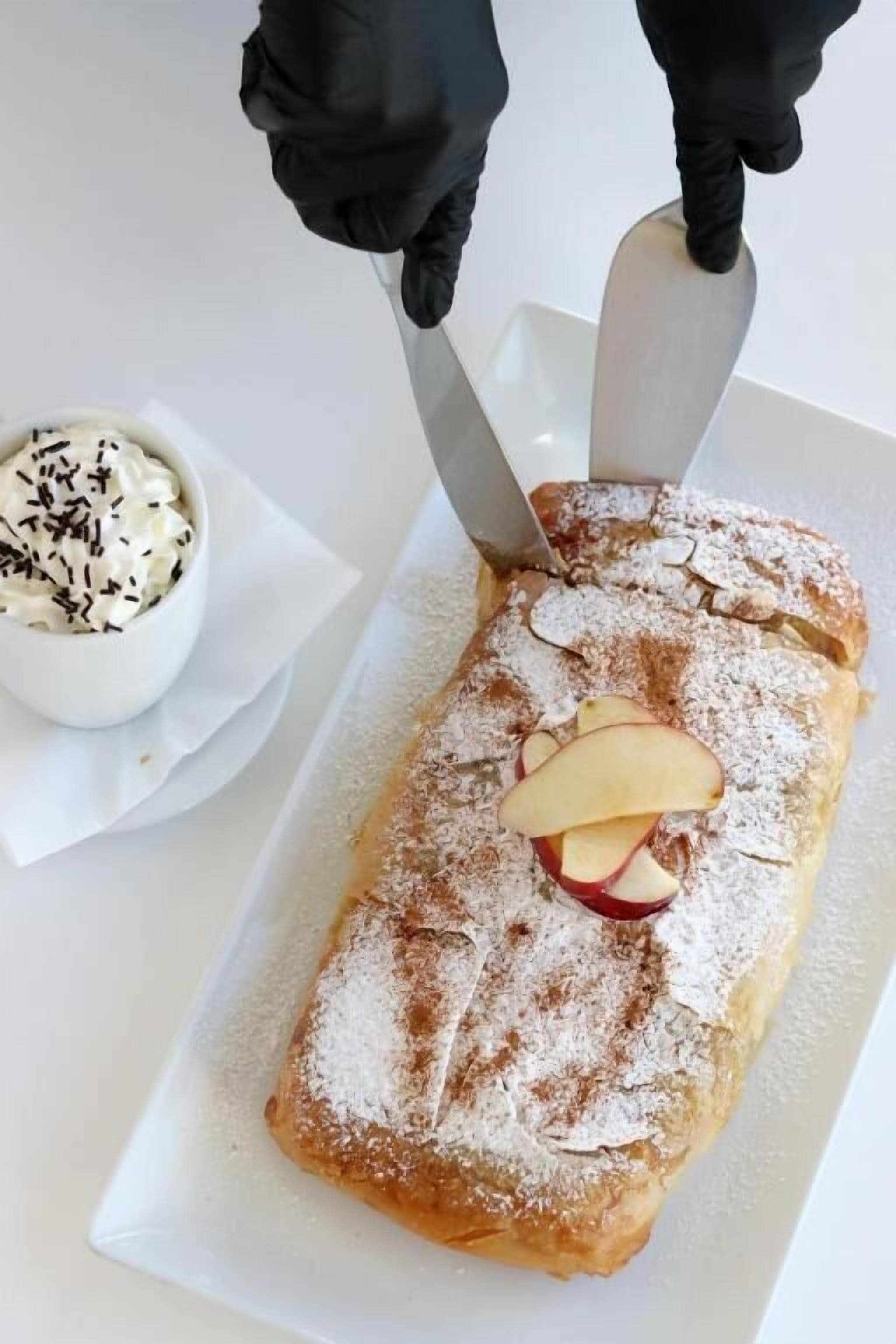 A rectangular pastry with powdered sugar and apple slices on top, being cut with a knife.